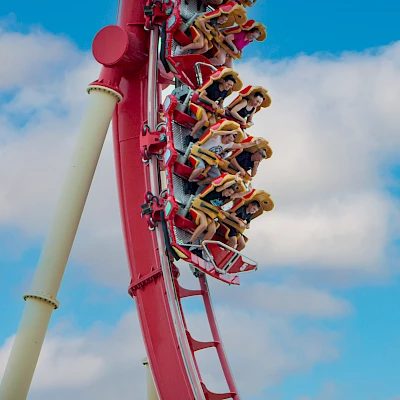 A group of people is enjoying a thrilling ride on a red roller coaster, captured mid-loop against a backdrop of a clear blue sky with some clouds.