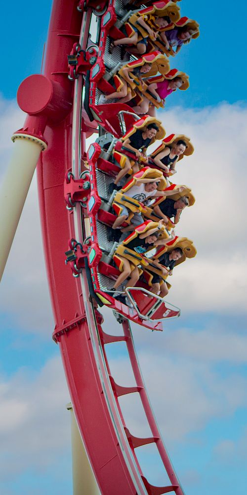 A group of people is enjoying a thrilling ride on a red roller coaster, captured mid-loop against a backdrop of a clear blue sky with some clouds.