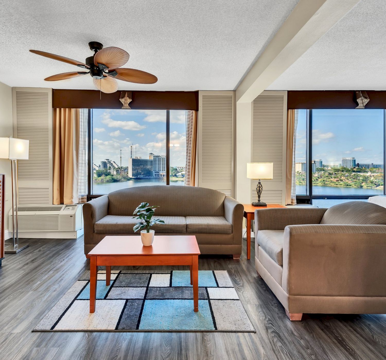 A modern hotel room features seating, decor, a ceiling fan, bed, and large windows with city views, creating a comfortable and inviting space.