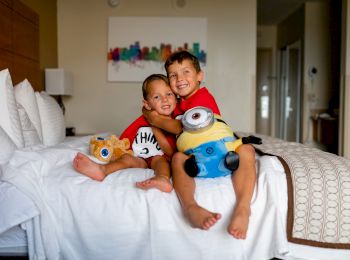 Two children are sitting on a bed in a room, each hugging a stuffed toy, while smiling at the camera.
