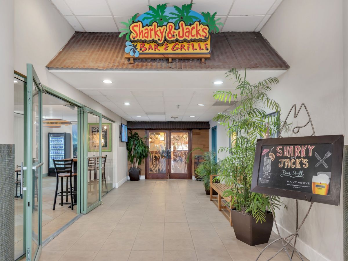 The image shows the entrance to "Sharky & Jack's Bar & Grille," with a sign and plants. There's a menu board and open glass door on the left ending the sentence.
