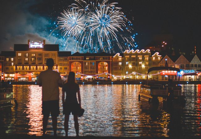 A couple stands near a waterfront, watching fireworks illuminate the night sky, with brightly lit buildings in the background.