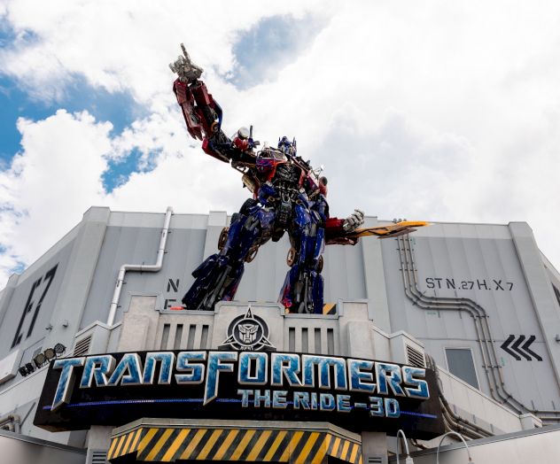 A large robot statue stands atop a building with the sign "Transformers The Ride 3D" against a partly cloudy sky.