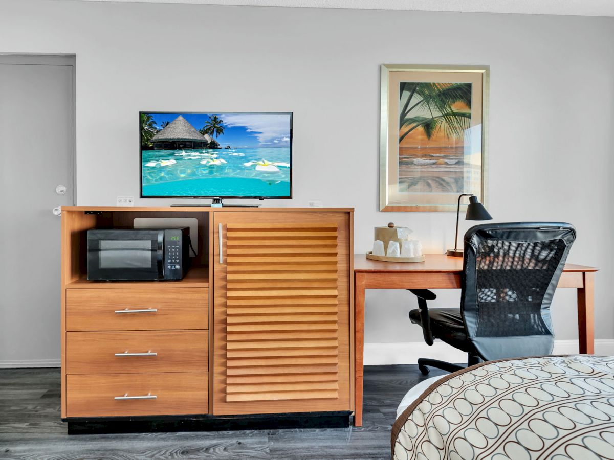 A hotel room with a desk, office chair, wall art, TV on a wooden dresser with a microwave, and part of a bed visible.
