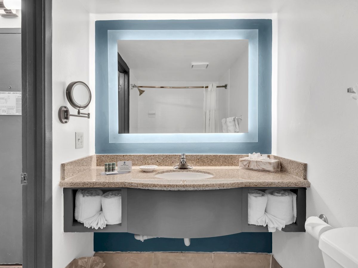 A modern bathroom featuring a lighted mirror above a sink with countertop space, towels, and toiletries, adjacent to a visible shower area.