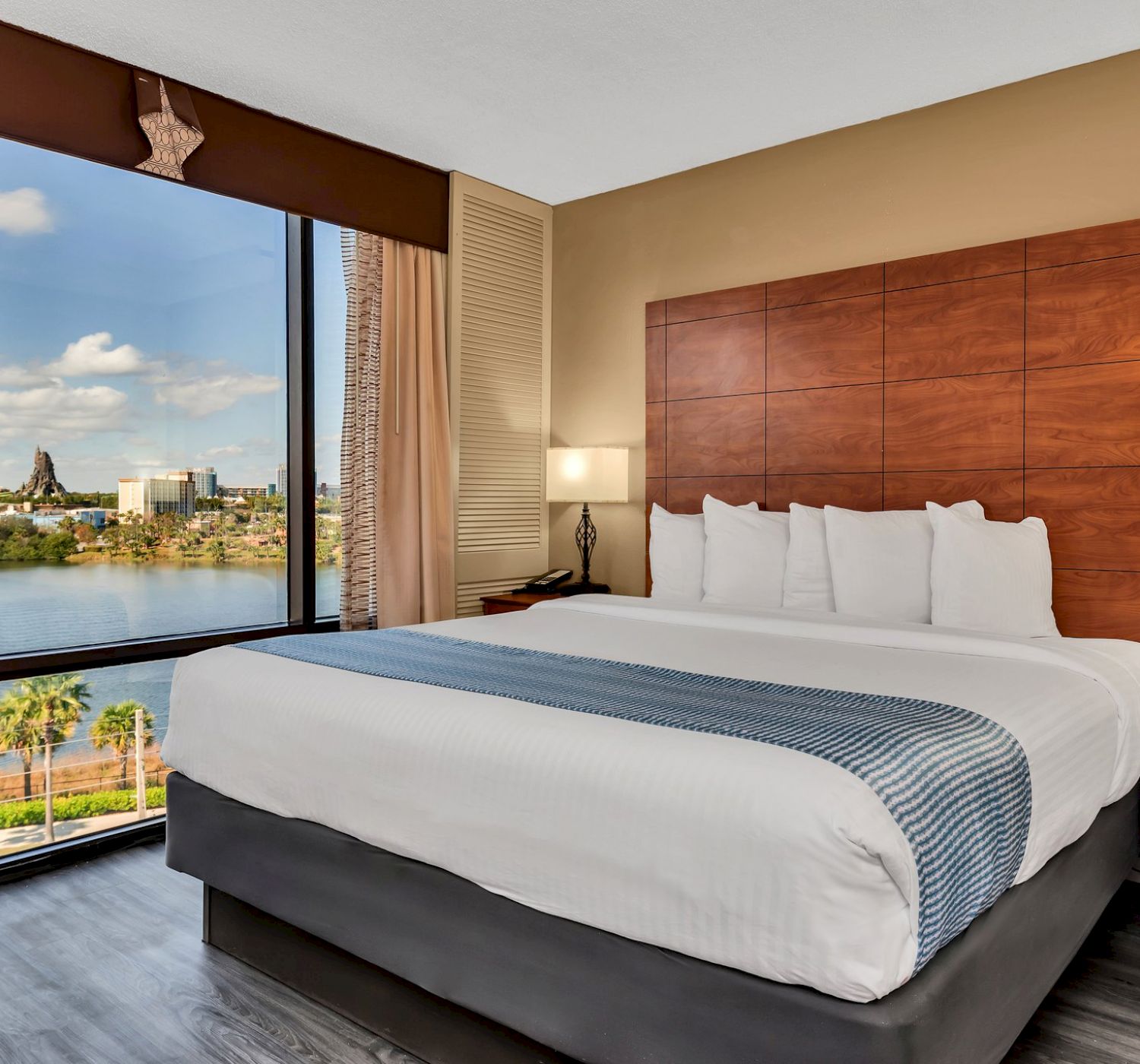 A hotel room with a large bed, bedside tables with lamps, and a view of the cityscape and lake through a wide window.