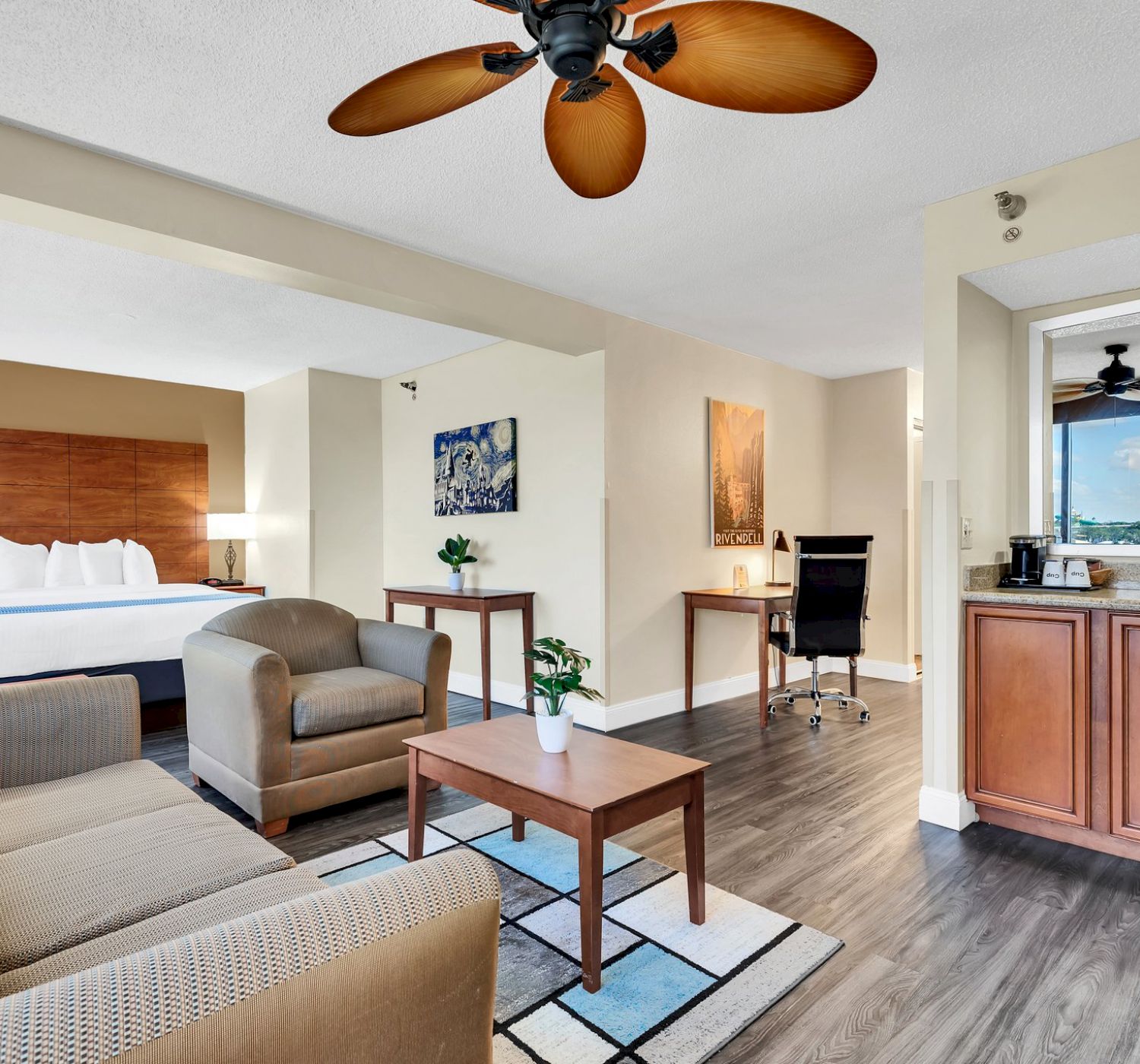 This image shows a modern hotel room with a bed, a sitting area, a work desk, a kitchenette, and a ceiling fan.