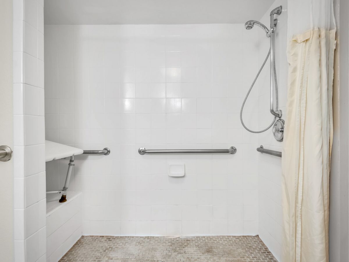 The image shows an accessible shower with a foldable bench, grab bars, a handheld showerhead, a tiled floor, and a curtain on the right side.