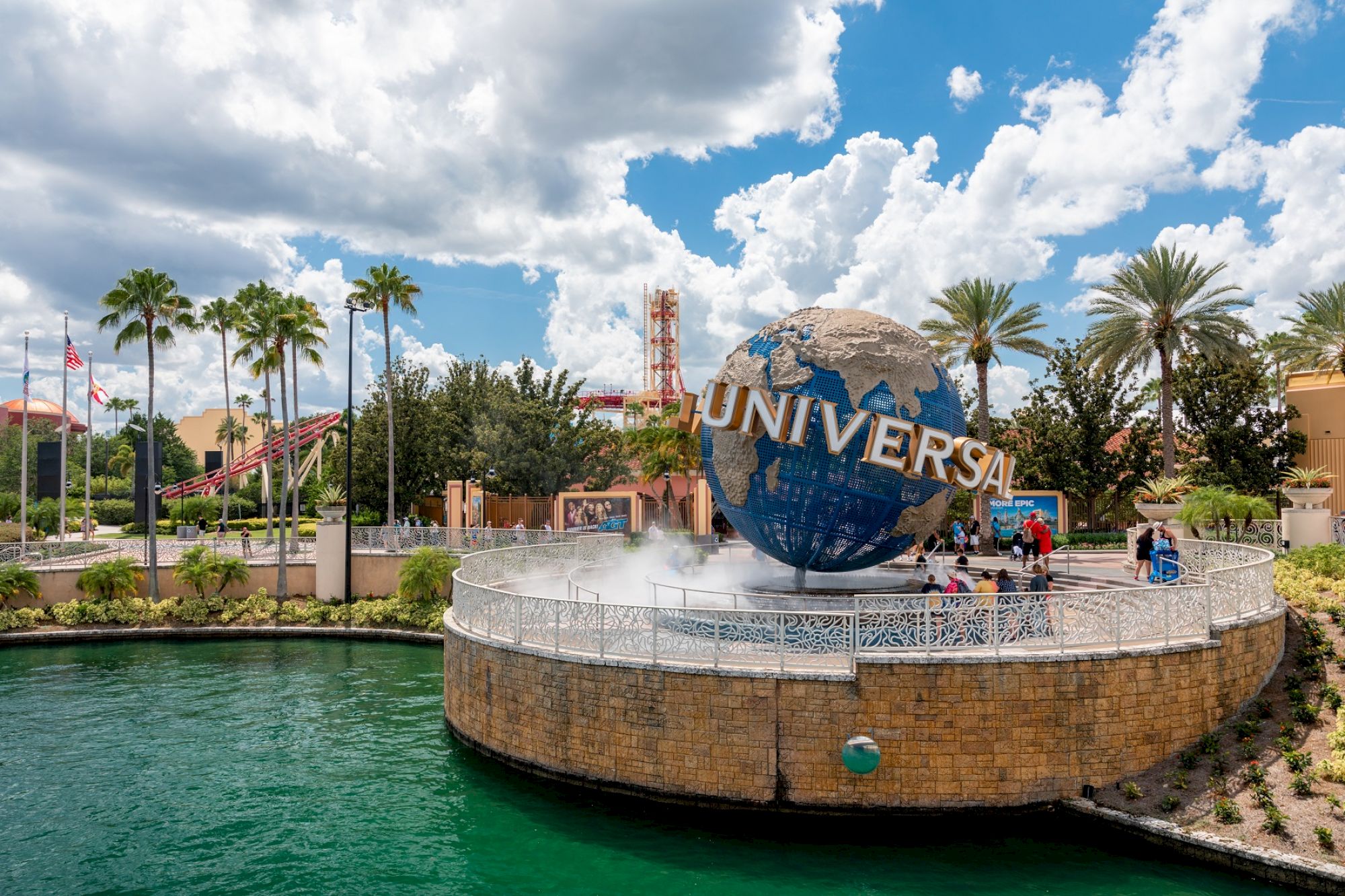 An amusement park scene with a large globe structure prominently displaying "Universal." Palm trees and water are visible in the foreground.