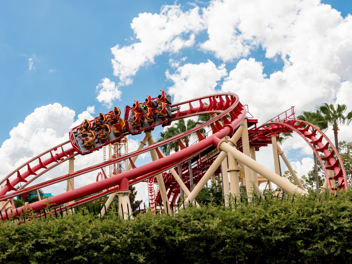 A red rollercoaster with passengers rides through a loop under a partly cloudy sky, with greenery in the foreground.