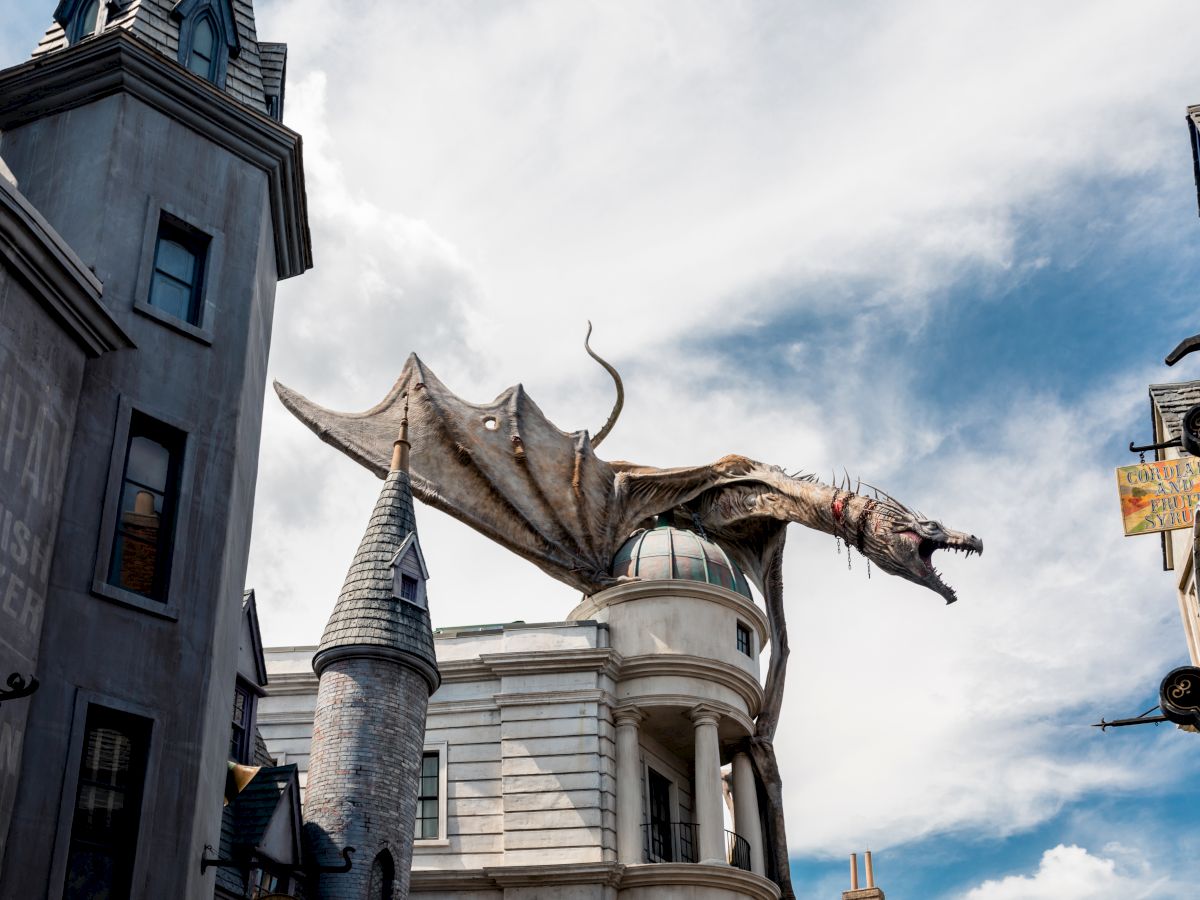 A large, detailed dragon sculpture is perched atop a building in a themed amusement park, with towers and a cloudy sky as the backdrop.