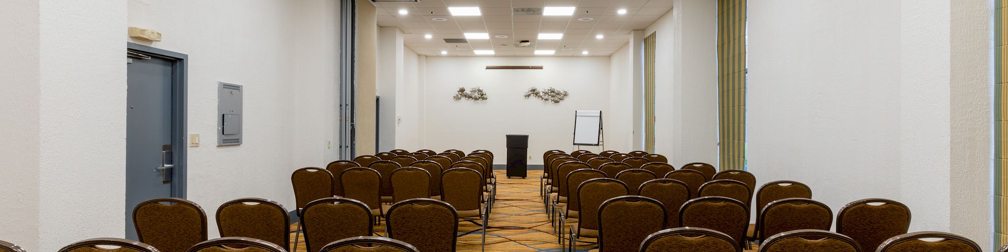The image shows a conference room with rows of chairs facing a podium, whiteboard, and a decorated wall. The room has a carpeted floor.