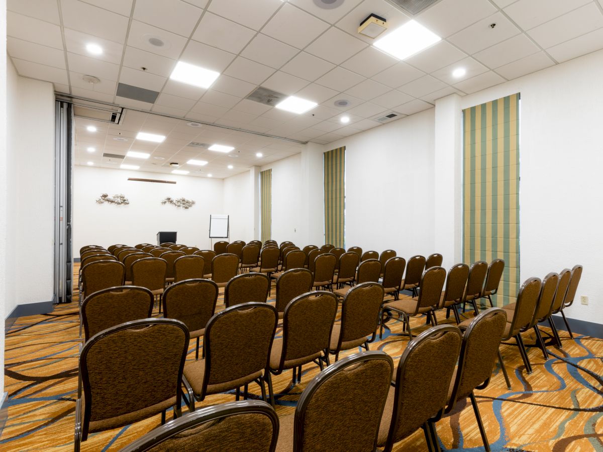 A conference room with rows of empty chairs arranged facing a screen or whiteboard, with patterned carpeting and neutral-colored walls.