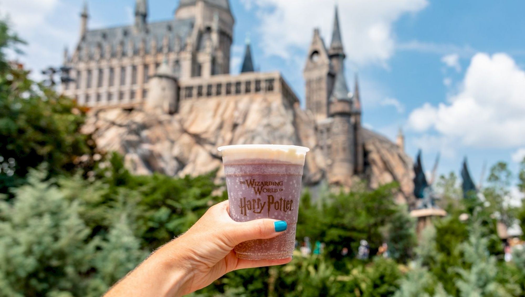A hand holding a "Wizarding World of Harry Potter" branded drink, with a castle resembling Hogwarts in the background on a sunny day.