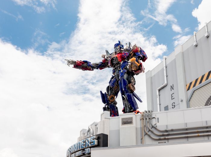 The image shows a large robotic figure resembling Optimus Prime from Transformers, standing on top of a building with "TRANSFORMERS" signage.