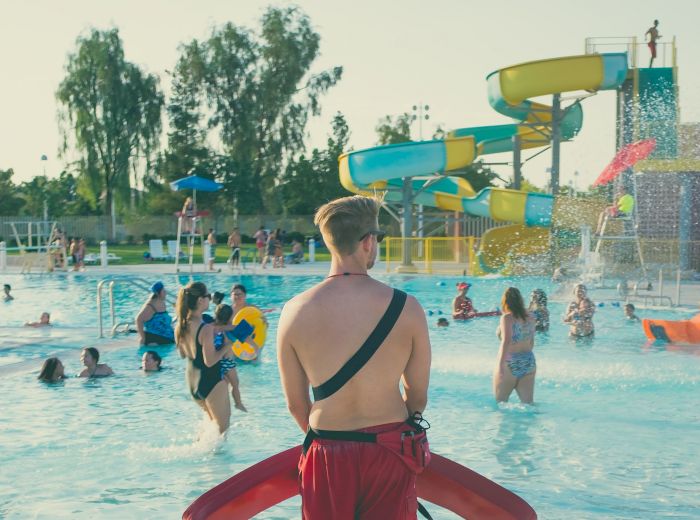 A lifeguard watches over a busy public pool with swimmers enjoying the water and various slides, on a warm, sunny day.