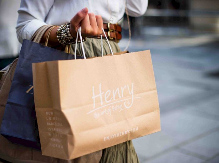 A person is holding a brown shopping bag with the name "Henry" on it, along with additional bags, while wearing a white shirt and bracelets.