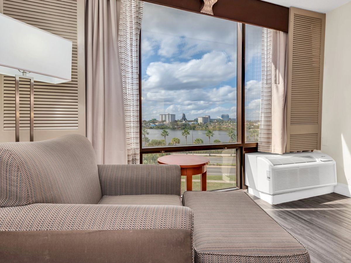 A room with a couch, floor lamp, large window with city view, small round table, and an air conditioner on the floor.