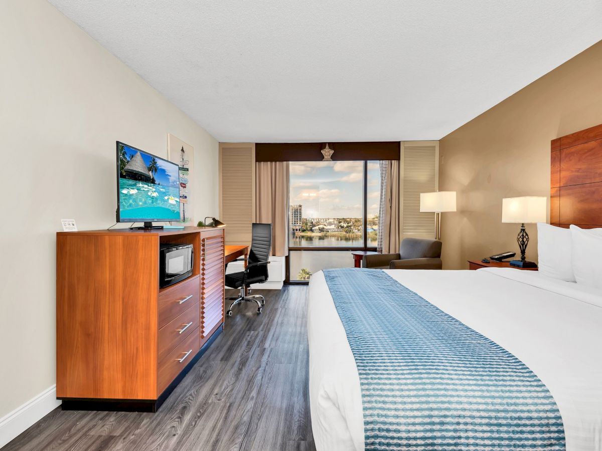 The image shows a modern hotel room with a king-size bed, a flat-screen TV, a desk, and a window with a view outside, featuring clean decor.
