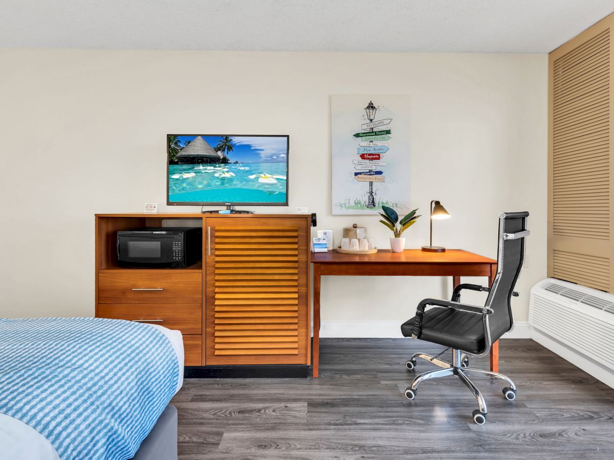 A neatly arranged hotel room with a bed, a desk with a chair and a lamp, a cabinet with a microwave, and a TV displaying a beach scene ending the sentence.
