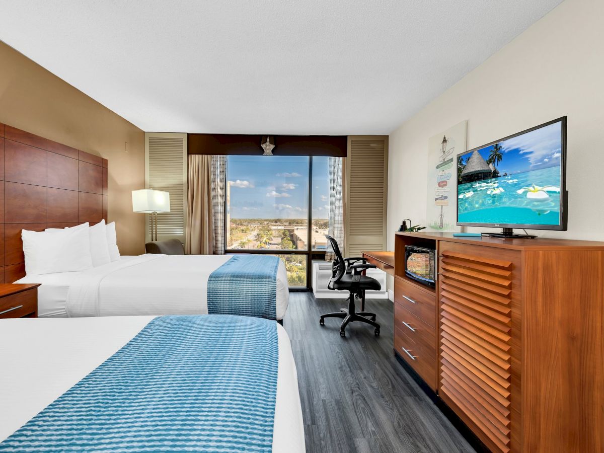 A hotel room with two double beds, a TV, a desk with a chair, and a large window with a view outside. The decor is modern and clean.