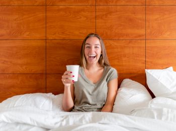 A woman is sitting up in bed, smiling, and holding a white cup, with a wooden headboard behind her and white pillows around.
